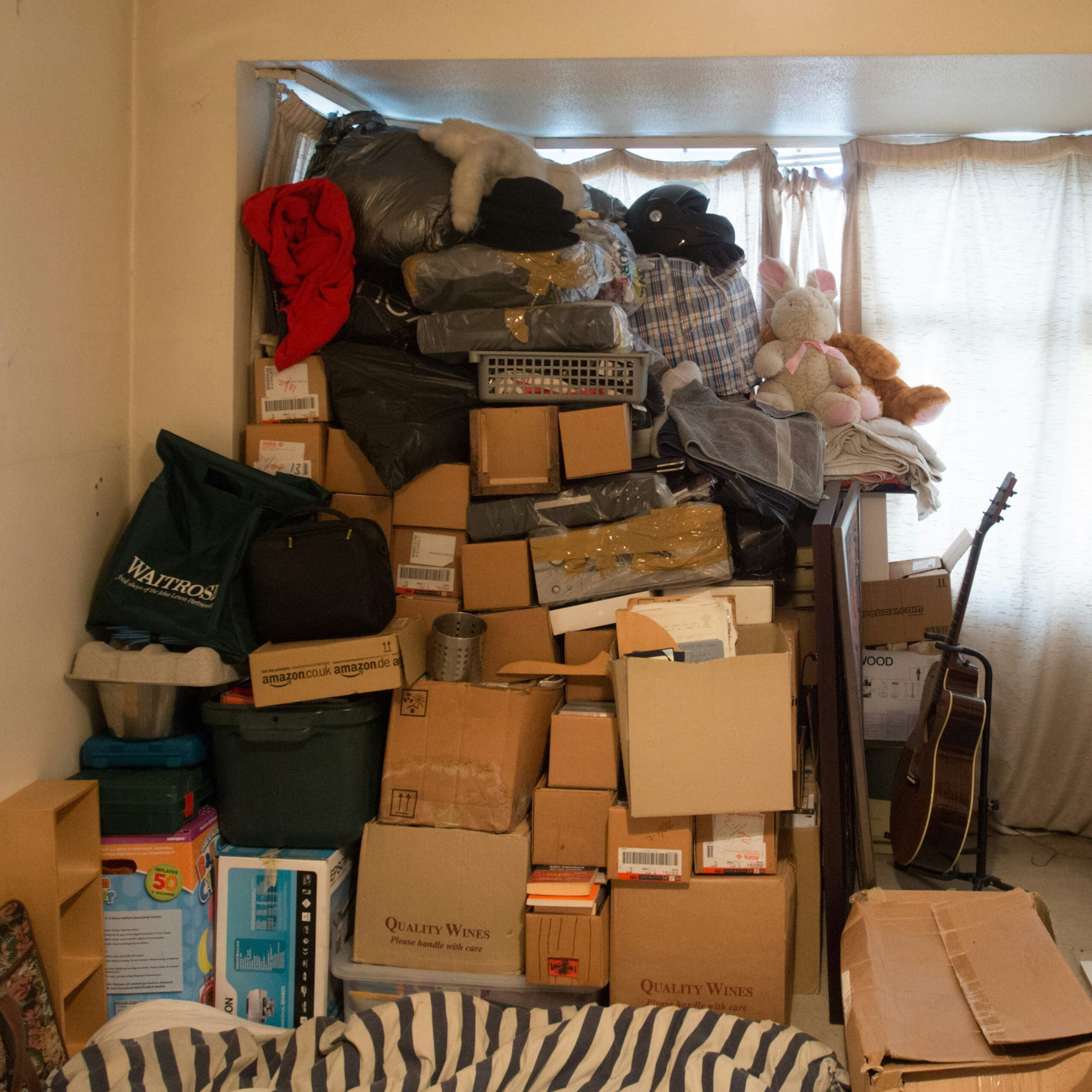 Alcôve encombrée de boîtes d'emballage (Quality Wines, Amazon), sacs, linge, et deux lapins en peluche près d'une fenêtre lumineuse.