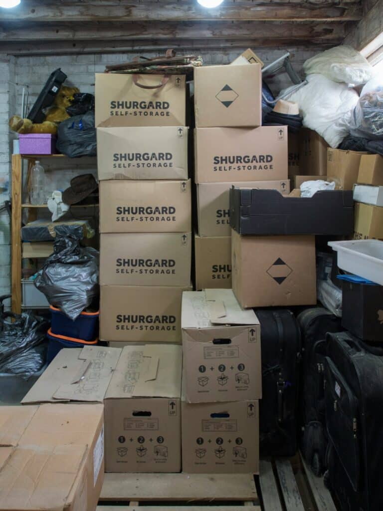 Shurgard : Box de stockage saturé, cartons et objets divers Piles de cartons de déménagement SHURGARD dans un box de stockage encombré, avec valises noires, sacs et divers objets stockés sur des étagères.